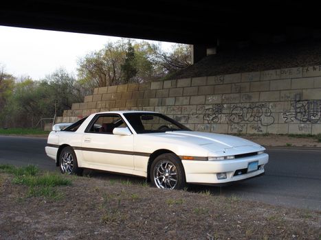 a shot of a white japanese sports car underneath a graffiti covered highway overpass.