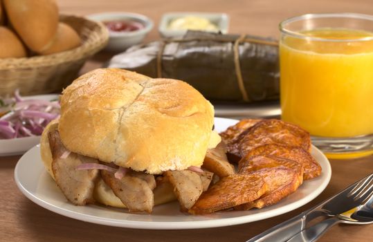 Typical Peruvian breakfast consisting of Pan con Chicharron (Bun with fried meat) and fried sweet potato, tamal (in the back), salsa criolla (onion salad) with orange juice and buns (Selective Focus, Focus on the front)