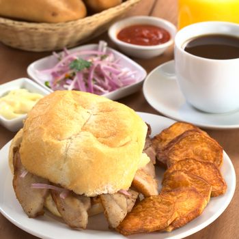 Typical Peruvian breakfast consisting of Pan con Chicharron (Bun with fried meat) and fried sweet potato, salsa criolla (onion salad), ketchup, mayonnaise, with coffee, orange juice and buns (Selective Focus, Focus on the front of the bun and the meat)