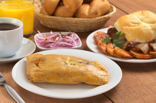 Peruvian breakfast with tamal (cooked corn with meat) and pan con chicharron (bun with fried pork meat), salsa criolla (onion salad), coffee, orange juice and bread (Selective Focus, Focus on the front of the tamal)