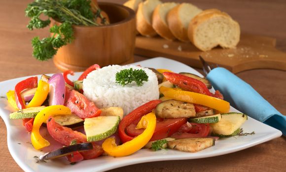 Ratatouille of zucchini, eggplant, tomato, bell pepper and onion with cooked rice and mortar with herbs and baguette in the back (Selective Focus, Focus on the front of the rice and the vegetable around)