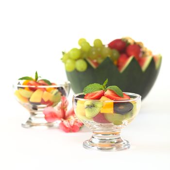 Fresh fruit salad with strawberry, kiwi, mango and grapes in glass bowl garnished with mint leaf with Inca Lily and a watermelon bowl full of fruits in the back (Selective Focus, Focus on the mint leaf in the first bowl)