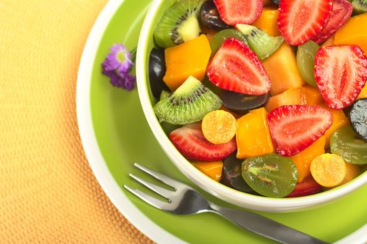 Fresh and healthy fruit salad with strawberry, kiwi, grape, mango and physalis in a bowl with fork and small blue flowers photographed from above (Selective Focus, Focus from the front to the middle of the bowl)