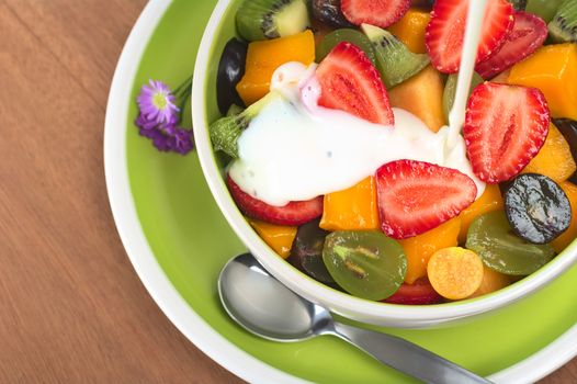 Fresh and healthy fruit salad with strawberry, kiwi, grape, mango and physalis in a bowl with plain yoghurt being poured over 