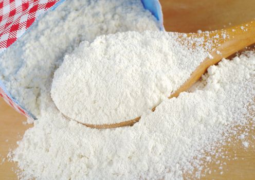 Wheat flour on wooden spoon lying in front of the paper package (Selective Focus, Focus on the flour on the spoon)