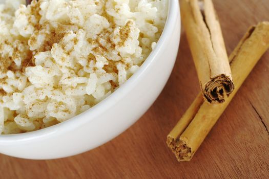 Rice pudding with ground cinnamon on top and cinnamon sticks on the side on wood (Selective Focus, Focus on the front rim of the bowl and the rice in the front)