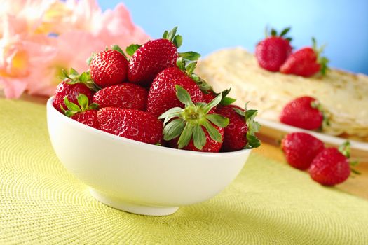 Fresh strawberries in white ceramic bowl with crepes and pink gladiolus flowers in the background (Selective Focus, Focus on the front of the strawberries)