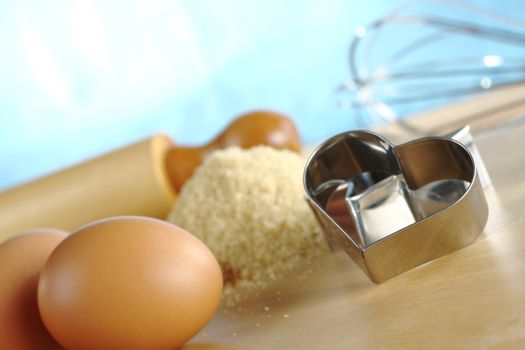 Heart shaped cookie cutter with baking ingredients and utensils such as eggs, brown sugar, rolling pin and a beater (Selective Focus, Focus on the front of the cookie cutter)