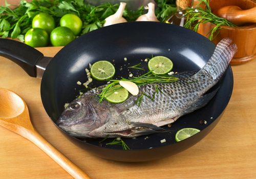 Raw tilapia with condiments (pepper corns, lime slices, garlic and rosemary) in frying pan with a wooden stirring spoon (Selective Focus, Focus on the head and belly of the fish)