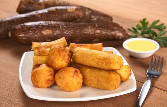 Filled fried balls and sticks out of manioc with a dip and some manioc roots in the back (Selective Focus, Focus on the manioc balls and the manioc sticks in the front)