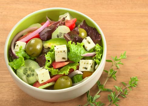 Greek salad out of cheese, green olives, tomato, green bell pepper, red onion, cucumber and lettuce with fresh oregano beside the bowl on wood (Selective Focus, Focus from the front to the middle of the bowl) 