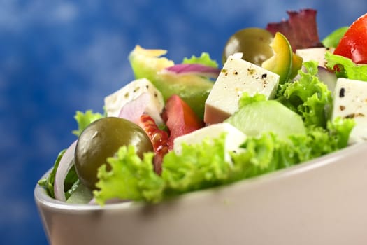 Greek salad out of cheese, green olives, tomato, green bell pepper, red onion, cucumber and lettuce with blue background (Selective Focus, Focus on the cheese in the middle) 