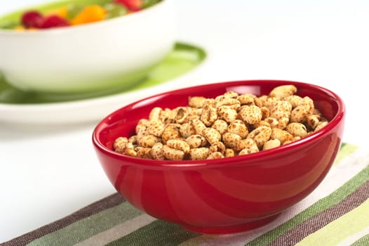 Puffed wheat cereal in red bowl with fruit salad in the background (Selective Focus, Focus on the middle of the cereals)