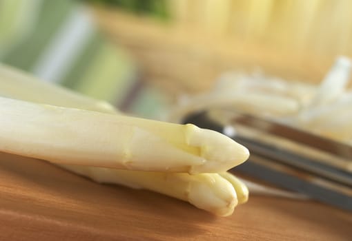 Two white asparagus with peeler and asparagus peel in the back on wooden board (Very Shallow Depth of Field, Focus on the two asparagus heads)