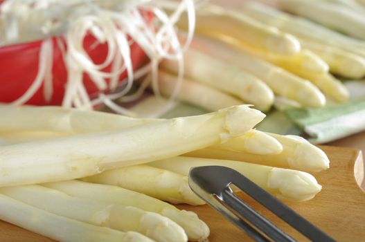 White asparagus with peeler on wooden board with asparagus and a red bowl full of peel in the back (Selective Focus, Focus on the asparagus lying on top) 