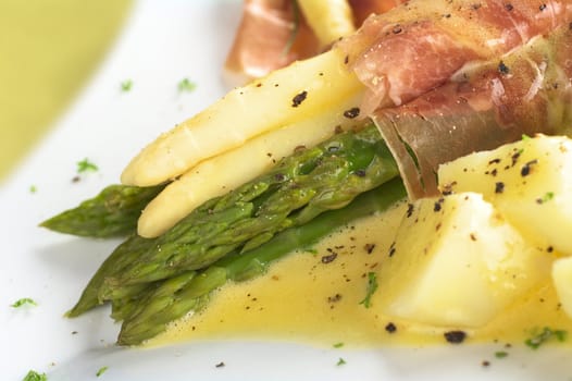 White and green asparagus wrapped in thin ham slices with Hollandaise sauce on top garnished with black pepper and parsley with cooked potato in front (Selective Focus, Focus on the front of the green asparagus and ham) 