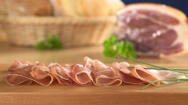 Thin ham slices on cutting board garnished with chives with piece of ham and bread basket in the back (Very Shallow Depth of Field, Focus on the front of the ham slices)