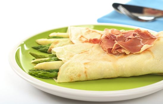 Green and white asparagus wrapped in crepes with ham as garnish on top (Selective Focus, Focus on the asparagus in the front and part of the ham)