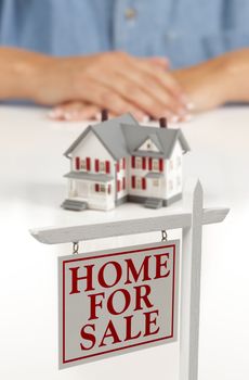 Womans Folded Hands Behind Model House and Home For Sale Real Estate Sign In Front on White Surface.