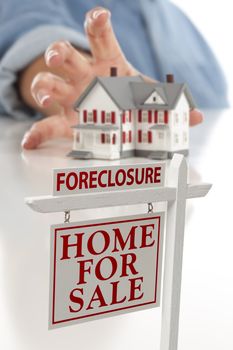 Foreclosure Real Estate Sign in Front of Womans Hand Reaching for Model House on a White Surface.