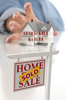 Sold Real Estate Sign in Front of Womans Hand Reaching for Model House on a White Surface.