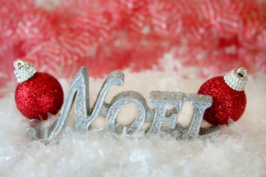 The word Noel made of glitter sitting in the middle of fake snow with two red christmas ornaments on each end.  Used selective focus and a shallow depth of field.