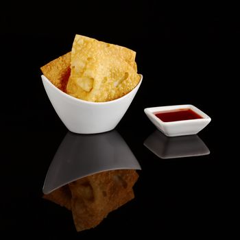 Chinese fried wantan chips in a white bowl with a sweet red sauce on black 