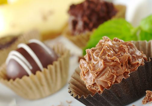 Chocolate truffles with mint leaf, fine chocolate shavings and a yellow cake in the background (Very Shallow Depth of Field, Focus on the front of the first praline) 