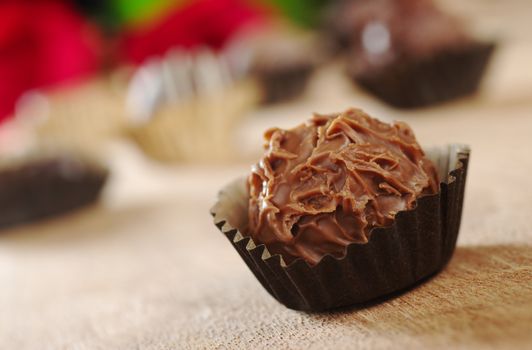Closeup of a milk chocolate truffle with wrapping, with other truffles and red roses in the background photographed on wood (Very Shallow Depth of Field, Focus on the front of the first praline) 