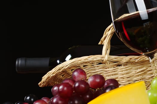 Red wine bottle in basket with wine glass, grapes and cheese in front (Selective Focus, Focus on the wine bottle)