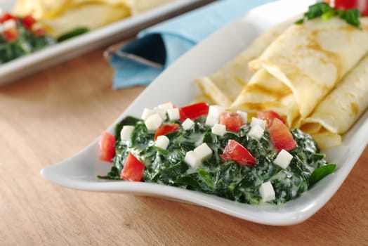 Spinach-tomato-cheese sauce with pancakes on a long plate with cutlery wrapped in blue napkin on wood (Selective Focus, Focus on the front of the sauce)