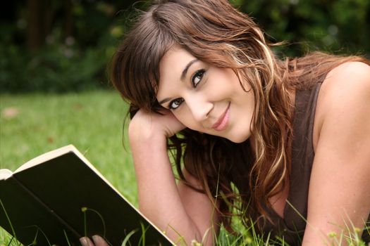 Pretty young woman laying on green grass and reading a book