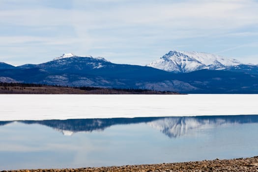 First open water at thawing Lake Laberge, Yukon Territory, Canada: reflection of snow-covered mountains on calm open water surface of still largely ice-covered lake.