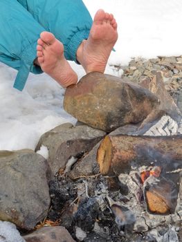 Bare feet of one person are warming at a Campfire outdoors at wintertime with patches of snow.
