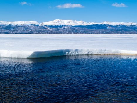 First open water at thawing Lake Laberge, Yukon Territory, Canada, and snow-covered mountains at distant shore of ice-covered frozen lake.