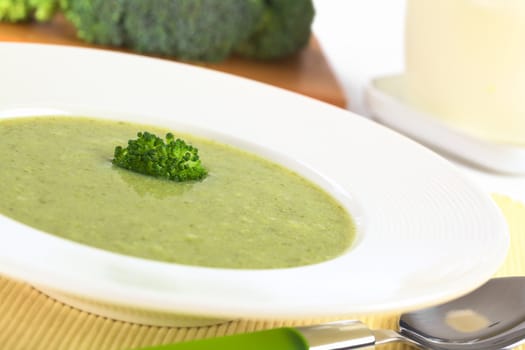 Cream of broccoli garnished with a broccoli floret on top with milk and raw broccoli in the back (Selective Focus, Focus on the broccoli floret on the soup)