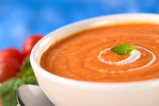 Cream of tomato with a small spiral of cream on top and a basil leaf with tomatoes and basil leaves in the back (Selective Focus, Focus on the basil leaf in the bowl)