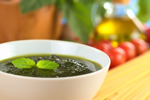 Fresh pesto made of basil, garlic and olive oil in a bowl and garnished with a basil leaf with raw pasta, cherry tomatoes, olive oil and mortar in the back (Selective Focus, Focus on the basil leaf in the bowl)