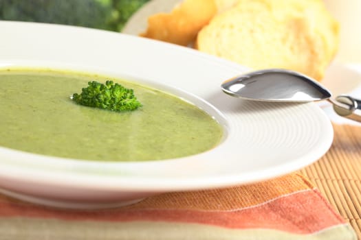 Cream of broccoli garnished with a broccoli floret on top with a spoon on the side of the plate and with baguette slices and raw broccoli in the back (Selective Focus, Focus on the broccoli floret on the soup)