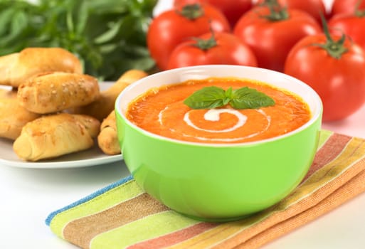 Cream of tomato with a spiral of cream on top and bread, tomatoes and basil leaves in the back (Selective Focus, Focus on the basil leaf in the bowl)