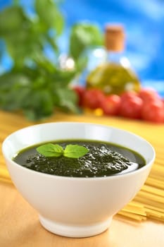 Fresh pesto made of basil, garlic and olive oil in a bowl and garnished with a basil leaf with raw pasta, cherry tomatoes, olive oil and basil leaves in the back (Selective Focus, Focus on the basil leaf in the bowl)