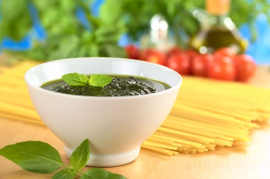 Fresh pesto made of basil, garlic and olive oil in a bowl and garnished with a basil leaf with raw pasta, cherry tomatoes, olive oil and basil leaves in the back (Selective Focus, Focus on the basil leaf in the bowl)
