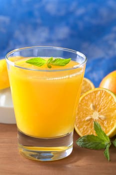 Freshly squeezed orange juice with orange slice and mint leaf on top of the juice with a blue background (Selective Focus, Focus on the mint leaf on top of the juice)