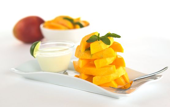 A pile of mango sticks garnished with mint leaf with a bowl of yogurt dip in the back (Selective Focus, Focus on the front of the pile)