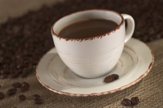 Hot fresh coffee in cup surrounded by coffee beans on fabric called Jute (Very Shallow Depth of Field, Focus on the front of the rim)