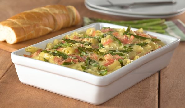 Casserole of green asparagus, ham and macaroni with baguette slices and plates in the back (Selective Focus, Focus in the middle of the dish)