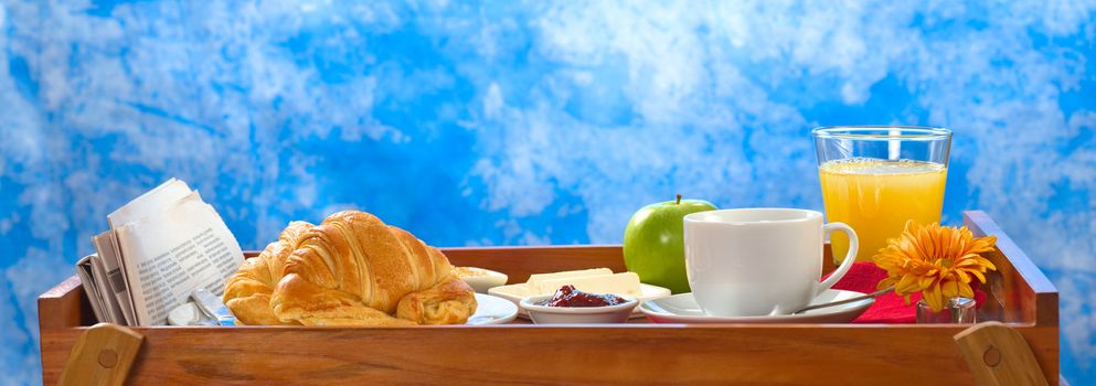 Delicious breakfast on tray consisting of coffee, orange juice, apple, croissants, jam and butter with newspaper lying on the side
