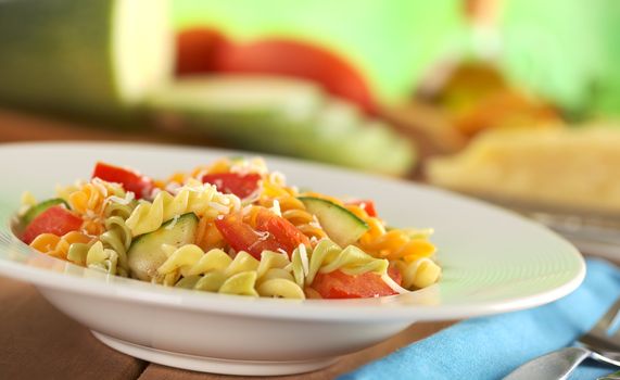 Colorful fusilli pasta with zucchini, tomato and grated cheese with fork on the side and ingredients in the back (Selective Focus, Focus one third into the meal)