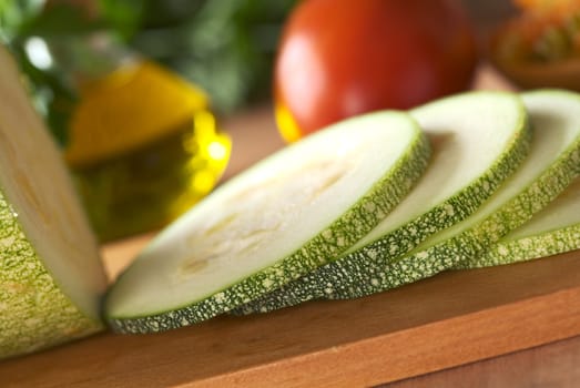 Zucchini slices on cutting board with tomato, oil and parsley in the back (Very Shallow Depth of Field, Focus on the front of the slices)  