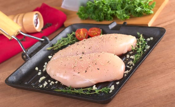 Raw chicken breast in frying pan seasoned with pepper, garlic and thyme with fresh herb as well as a pepper mill in the back (Selective Focus, Focus on the front of the meat)